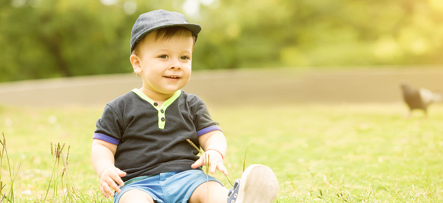 Smiling toddler wearing a cap and casual clothes, sitting on the grass in a sunny park