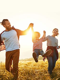 Happy family of three walking in a grassy field at sunset, with the parents swinging their child between them