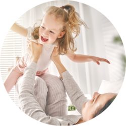 Mother and young daughter playing together, with the mother lifting the laughing child into the air, in a bright room with light filtering through the blinds.